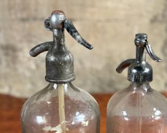 Vintage soda siphons featuring glass bodies and metal dispensers. One bottle has visible etched lettering, indicating it is from St. Louis, Missouri. Both show signs of wear and age, including discoloration, patina on the metal tops, and residue inside. These pieces are suitable as decorative or collectible items.