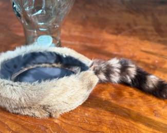 A fur cap featuring a raccoon tail, displayed on a handmade glass mannequin head. The cap’s material is natural fur, with a thick soft lining for comfort. The glass head is crafted from recycled glass and made in Spain. Both the cap and glass head appear in good condition.