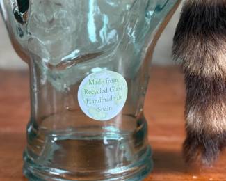A fur cap featuring a raccoon tail, displayed on a handmade glass mannequin head. The cap’s material is natural fur, with a thick soft lining for comfort. The glass head is crafted from recycled glass and made in Spain. Both the cap and glass head appear in good condition.