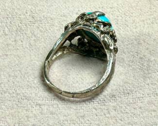 Sterling silver ring featuring a vibrant turquoise cabochon centerpiece. The band is ornately designed with detailed organic elements resembling leaves or vines. The turquoise stone is prominently set, securely held by prongs. The interior of the band shows slight wear, consistent with age and previous use. Comes in a size 6.