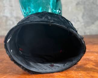 Black faux fur hat with a circular design, displayed on a translucent green glass head made from recycled materials and handcrafted in Spain. The interior of the hat is lined with fabric and features small red details. The glass head has a textured surface with a visible sticker stating its recycled origin.