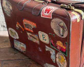 Brown leather suitcase adorned with various vintage travel decals from international destinations. Features brass hardware, interior cloth lining, and built-in hangers. Includes a monogram ‘L.M.’ on the leather exterior. Condition shows wear consistent with use, including scuffs and scratches on the leather and minor handling marks inside. No key.