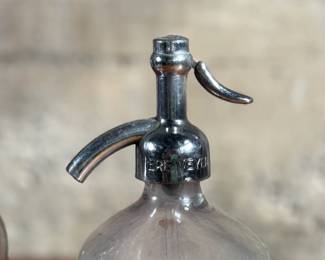 Vintage soda siphons featuring glass bodies and metal dispensers. One bottle has visible etched lettering, indicating it is from St. Louis, Missouri. Both show signs of wear and age, including discoloration, patina on the metal tops, and residue inside. These pieces are suitable as decorative or collectible items.