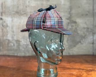 Tartan deerstalker cap featuring a classic red, blue, and white plaid pattern with a black ribbon detail on top. Inner lining crafted from black fabric for added comfort, and a label marked “Tartan Gift Shop by Lynton” is present. Displayed on a handblown glass head form, made from recycled glass in Spain, as indicated by the sticker. A distinctive decorative or collectible piece.
