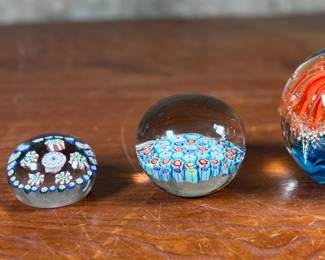 Three glass paperweights featuring intricate floral and abstract designs. Includes one large piece with orange floral motifs and two smaller pieces showcasing millefiori patterns in vibrant colors. These paperweights are in excellent condition with no visible chips or cracks. No visible signatures seen.