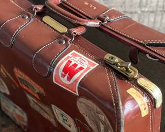 Brown leather suitcase adorned with various vintage travel decals from international destinations. Features brass hardware, interior cloth lining, and built-in hangers. Includes a monogram ‘L.M.’ on the leather exterior. Condition shows wear consistent with use, including scuffs and scratches on the leather and minor handling marks inside. No key.