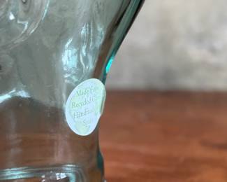 Tartan deerstalker cap featuring a classic red, blue, and white plaid pattern with a black ribbon detail on top. Inner lining crafted from black fabric for added comfort, and a label marked “Tartan Gift Shop by Lynton” is present. Displayed on a handblown glass head form, made from recycled glass in Spain, as indicated by the sticker. A distinctive decorative or collectible piece.