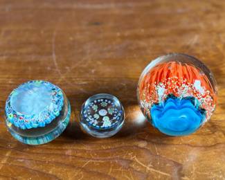 Three glass paperweights featuring intricate floral and abstract designs. Includes one large piece with orange floral motifs and two smaller pieces showcasing millefiori patterns in vibrant colors. These paperweights are in excellent condition with no visible chips or cracks. No visible signatures seen.