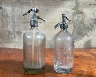 Vintage soda siphons featuring glass bodies and metal dispensers. One bottle has visible etched lettering, indicating it is from St. Louis, Missouri. Both show signs of wear and age, including discoloration, patina on the metal tops, and residue inside. These pieces are suitable as decorative or collectible items.