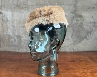 A fur cap featuring a raccoon tail, displayed on a handmade glass mannequin head. The cap’s material is natural fur, with a thick soft lining for comfort. The glass head is crafted from recycled glass and made in Spain. Both the cap and glass head appear in good condition.