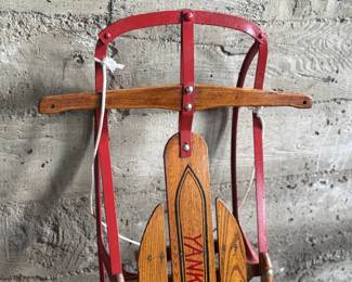 Wooden Flexible Flyer Yankee Clipper sled featuring steel runners painted in red and a wooden deck with printed branding. The sled includes a wooden crossbar for steering and a rope control. Signs of wear consistent with age and use, including scratches and minor rust on metal parts, are visible. A classic item for collectors or enthusiasts of vintage winter gear. Measures, 44” H x 20.5” W.
