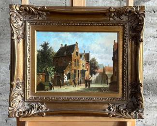 Framed oil on panel painting depicting a detailed village scene with several figures and buildings. Signed by the artist in the lower right corner. Encased in an ornate, gilt wooden frame with floral details. Measures in frame, 20.75” H x 24.75” W.