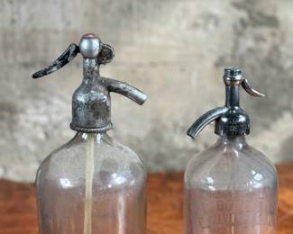 Vintage soda siphons featuring glass bodies and metal dispensers. One bottle has visible etched lettering, indicating it is from St. Louis, Missouri. Both show signs of wear and age, including discoloration, patina on the metal tops, and residue inside. These pieces are suitable as decorative or collectible items.