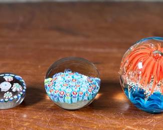Three glass paperweights featuring intricate floral and abstract designs. Includes one large piece with orange floral motifs and two smaller pieces showcasing millefiori patterns in vibrant colors. These paperweights are in excellent condition with no visible chips or cracks. No visible signatures seen.