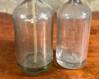 Vintage soda siphons featuring glass bodies and metal dispensers. One bottle has visible etched lettering, indicating it is from St. Louis, Missouri. Both show signs of wear and age, including discoloration, patina on the metal tops, and residue inside. These pieces are suitable as decorative or collectible items.