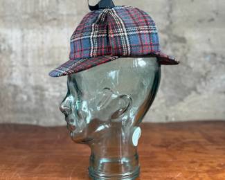 Tartan deerstalker cap featuring a classic red, blue, and white plaid pattern with a black ribbon detail on top. Inner lining crafted from black fabric for added comfort, and a label marked “Tartan Gift Shop by Lynton” is present. Displayed on a handblown glass head form, made from recycled glass in Spain, as indicated by the sticker. A distinctive decorative or collectible piece.