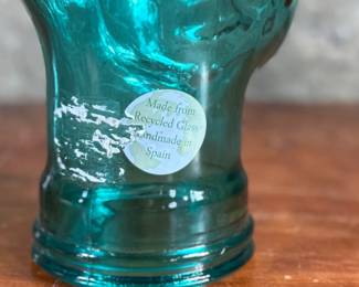 Black faux fur hat with a circular design, displayed on a translucent green glass head made from recycled materials and handcrafted in Spain. The interior of the hat is lined with fabric and features small red details. The glass head has a textured surface with a visible sticker stating its recycled origin.