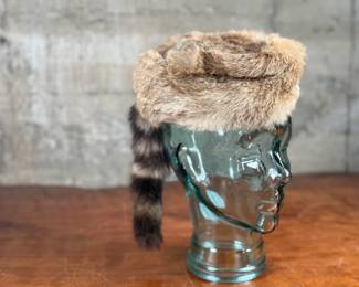 A fur cap featuring a raccoon tail, displayed on a handmade glass mannequin head. The cap’s material is natural fur, with a thick soft lining for comfort. The glass head is crafted from recycled glass and made in Spain. Both the cap and glass head appear in good condition.
