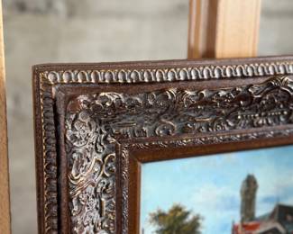 Framed oil painting depicting a European village scene featuring a bridge, architecture, and figures in a cobblestone street. The artwork comes in an ornately carved frame with intricate detailing. The reverse side is covered with paper backing and equipped with a hanging wire. Signed lower right by artist and measures in frame, 16” H x 18” W.