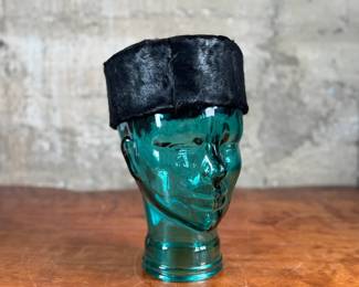 Black faux fur hat with a circular design, displayed on a translucent green glass head made from recycled materials and handcrafted in Spain. The interior of the hat is lined with fabric and features small red details. The glass head has a textured surface with a visible sticker stating its recycled origin.