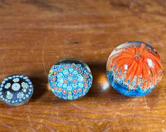 Three glass paperweights featuring intricate floral and abstract designs. Includes one large piece with orange floral motifs and two smaller pieces showcasing millefiori patterns in vibrant colors. These paperweights are in excellent condition with no visible chips or cracks. No visible signatures seen.