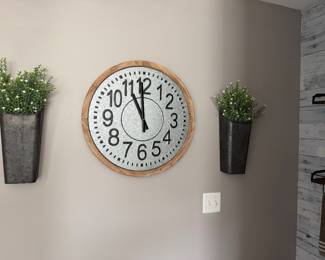 rustic farmhouse-style wall clock features a circular galvanized metal face with large black numerical markers and a light wood frame. 