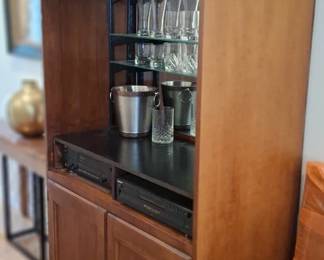 wooden bar cabinet resembles a cherry wood entertainment armoire repurposed with a mirrored back and glass shelves for displaying barware. doors removed for display are included