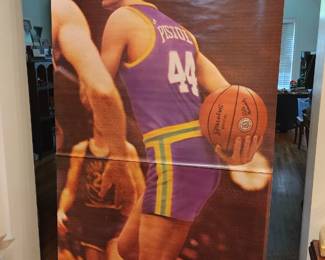 This vintage poster features Pete Maravich in a New Orleans Jazz uniform, dribbling a basketball while wearing a Spalding 6-foot-5 staff. Maravich, nicknamed "Pistol Pete," was a Hall of Fame scorer and playmaker who revolutionized the game in the 1960s and 1970s