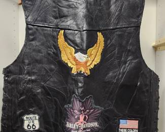 black leather biker vest adorned with multiple embroidered patches, including a golden eagle, a "Route 66" emblem, and a "Harley-Davidson" design. The vest also features a "Ride Free" patch and a "Loud Pipes Save Lives" sign, reflecting its association with motorcycle culture and road travel.