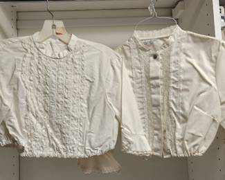 Two cream-colored vintage blouses hanging side by side, each featuring lace trim and button details. The garments appear to be from the mid-20th century