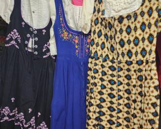 three vintage women's dresses hanging on a rack, each featuring unique patterns and embroidery. The garments display a mix of floral, geometric, and traditional designs, suggesting a collection of mid-20th-century or post-war fashion