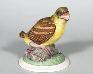 BOEHM (AMERICAN) BABY GOLDFINCH PORCELAIN FIGURINE, MODEL 448 | Bisque/porcelain polychrome figural of a juvenile goldfinch perched on a modeled tree-stump and floral base, hand-painted in yellow, brown, green and pink tones with an open beak and detailed feather modeling. Matte white circular base with a printed underside mark reading "Boehm / Baby Goldfinch / 448 I / Made in U.S.A." and a small vent hole. Features naturalistic glass-like eyes and finely incised feather texture. Overall, the piece is a detailed studio porcelain bird in the Boehm production style.