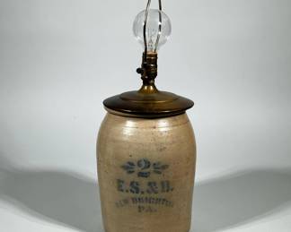 E.S. & B. STENCILED STONEWARE CROCK LAMP, NEW BRIGHTON PA | Converted stoneware crock lamp formed from a tan/beige, salt-glazed ovoid crock with a cobalt-blue stenciled numeral and leafy motif above the stenciled text "E.S. & B. NEW BRIGHTON PA." Fitted with a turned brass top, brass socket, harp and finial and shown with a clear bulb. Crock body shows typical kiln grit and small surface inclusions and the stencil lettering is visible on the front.