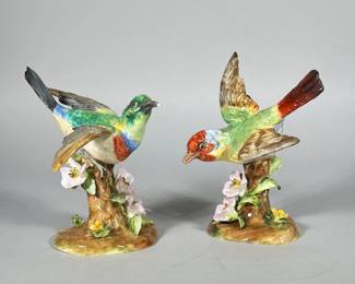 PAIR CROWN STAFFORDSHIRE FINE BONE CHINA BIRD FIGURINES | Pair of polychrome glazed fine bone china bird figural groups modelled perched on tree-stump bases with applied pink and yellow flowers. Hand-painted with green, blue, red, brown and cream enamels and finished with a high-gloss glaze; molded feather and floral detailing is evident. Bases feature molded root/grounding forms with applied leaves and blossoms and metal-styled legs securing the birds to the stumps. Dimensions: h. 6 x w. 4.5 x d. 3 in