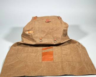 WAXED CANVAS WORK APRON WITH LEATHER STRAPS AND BRASS D-RINGS | Brown waxed canvas work apron with sewn pockets and leather-reinforced tool loops. Features adjustable leather buckle straps at the top, brass D-rings, rivet hardware and a front snap-closure strap. Large front flap pocket and several smaller sewn pockets including a narrow leather tool slot. Constructed with visible topstitching and a characteristic waxed finish.