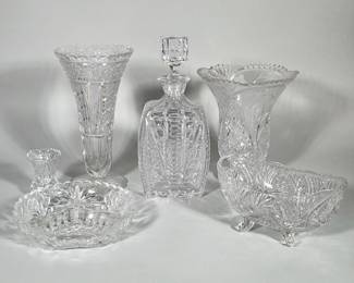 (6pc) BRILLIANT-STYLE CUT GLASS ASSORTED VASES, DECANTER AND BOWLS | Group of six clear brilliant-cut glass items including two tall trumpet vases, a faceted square-shouldered decanter with matching square stopper, a three-footed oval bowl, a shallow round bowl, and a small bud vase. Surfaces feature deep starburst, diamond/honeycomb and fan cuts with scalloped or beaded rims and molded/scroll feet on the footed bowl. The decanter is square-shouldered with a faceted square stopper; several pieces show complex intersecting panel cuts. All pieces are clear colorless glass or lead crystal with high-relief cutting and polished facets.