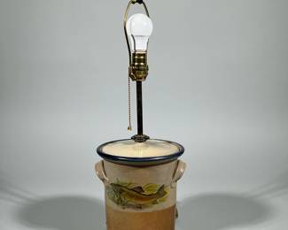 STONEWARE FISH MOTIF CROCK TABLE LAMP | Stoneware crock converted to a table lamp with fitted lid and brass harp, socket, and finial. Cylindrical two-tone body with natural tan-to-rust glaze, blue-rimmed lid, two loop handles and a painted or transfer fish motif on one side. Brass pull-chain socket and clear power cord installed through the base indicate a converted vessel used as lighting.