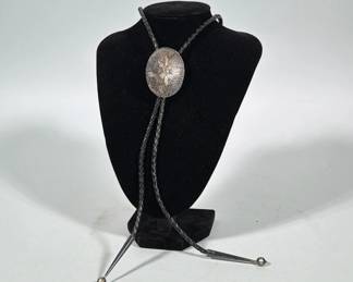 HIJE STERLING SILVER BOLO TIE WITH ENGRAVED OVAL SLIDE | Oval sterling slide with engraved radiating sunburst motif; verso stamped "STERLING" and "HIJE". Black braided leather cord with two tapered sterling tips terminated by small round ball finials. Slide secured to cord with a hinged metal clasp on the verso.