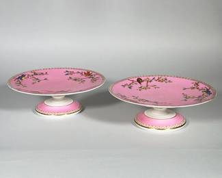 PAIR OF BODLEY PINK-GROUND PORCELAIN PEDESTAL COMPOTES | Pair of porcelain pedestal compotes with a pink enamel ground decorated with hand-painted polychrome birds and flowering branches. Rims and bases feature a gilt geometric border and gilt banding. Shallow circular dishes raised on turned pedestal bases with white glazed undersides. Decoration appears to combine transfer and hand-enamel painting.