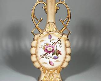 PORCELAIN URN WITH ENAMELED FLORAL PANELS AND GILT | Cream-ground porcelain urn with twin ornate gilt handles, narrow neck and molded ribbed sides. Front and back feature an oval polychrome enameled floral panel outlined in raised gold; additional raised gilt scrollwork at the foot and neck. Underside shows a drilled hole through the base and a blue painted script mark. Decoration appears applied with raised gilt outlines and hand-painted enamels in pink, purple and green tones.