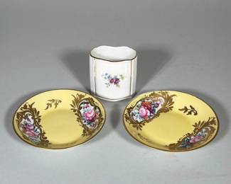 (3pc) LIMOGES HAND-PAINTED FLORAL SAUCERS WITH PLANKENHAMMER BAVARIA TOOTHPICK HOLDER | Two porcelain saucers with yellow ground, gilt rims and gilt foliate cartouche surrounds containing hand-painted pink and multicolor floral panels; underside of the saucers stamped "LIMOGES FRANCE" and inscribed in blue and red with hand-painting notation. Accompanying small white porcelain toothpick/toothpick holder with hand-painted floral spray and printed mark "Plankenhammer Floss Bavaria" and boxed "MADE IN GERMANY" on the base. Both saucers display overall gilt decoration and painted floral reserves; the toothpick holder has a molded rectangular form with a scalloped rim. Marks on the bases identify the saucers as Limoges-made with "décor main" (hand-decorated) and the small holder as Plankenhammer, Bavaria.