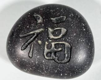 CARVED CHINESE 'FU' (?) STONE PEBBLE | Dark, oval polished pebble with light white speckling carved with the Chinese character ? (fu) in shallow relief on one face. The carved strokes appear slightly recessed and darker than the surrounding surface; the stone has a smooth, polished finish. Opposite face and underside are uncarved and similarly polished.