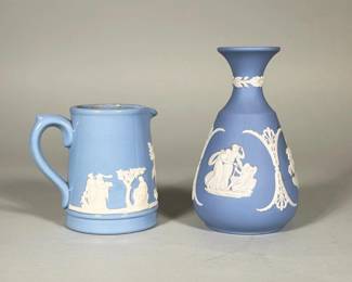 (2pc) WEDGWOOD JASPERWARE BLUE VASE AND CREAMER | Pair of pale-blue jasperware ceramic pieces consisting of a baluster bud vase and a small handled creamer, each decorated with applied white Neoclassical reliefs. One base shows a blue circular transfer mark reading "Jasper Ware Made in England Since 1800," the other base is impressed "WEDGWOOD" and "MADE IN ENGLAND" with mould numbers. The vase has a flared neck with a laurel band and oval classical medallions; the creamer features a continuous frieze, formed handle and a small spout. The creamer interior is glazed; both pieces show matte jasperware bodies with white applied ornament.