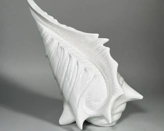WHITE GLAZED CERAMIC CONCH SHELL | White-glazed ceramic formed in the shape of a large conch shell with pronounced ribs and applied spines. Glossy white glaze covers the exterior with an open hollow interior accessed at the base. Underside shows an unglazed foot ring and a black ink inscription/mark. Surface displays molded naturalistic shell details including a flared lip and internal ridging.