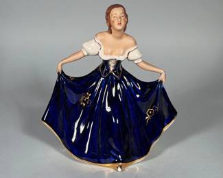 PORCELAIN DANCING LADY FIGURINE IN COBALT BLUE GOWN WITH GILT DECORATION | Porcelain figure of a standing woman holding the sides of a full cobalt-blue skirt with hand-applied gilt floral motifs and gilt trim at the hem. Modeled with exposed shoulders, molded hair and painted facial features in polychrome enamels. The interior and base show an impressed model number and a partial red circular factory/transfer mark; a small painted decorator's mark is visible on the underside. Glaze finish is glossy on the gown and more matte on the flesh areas.