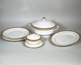(5pc) GLORIA (CZECHOSLOVAKIA) PORCELAIN SERVING PIECES WITH GILT RIM | Group of white porcelain service pieces with scalloped rims and a hand-applied gilt band. Includes a covered two-handled tureen with lid, a small covered sugar with saucer, two stacked dinner plates and a large oval platter. Underside backstamp shows a green shield with the word "GLORIA" and printed text reading "Czechoslovakia" and "leptaný dekor". Gilding shows an ornate etched pattern along the rim and gilt finials on the lids.
