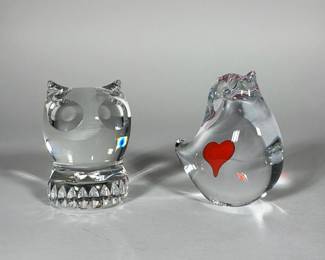 ART GLASS PAPERWEIGHT PAIR, SPODE OWL AND SIGNED HEART BIRD | Pair of clear glass animal paperweights comprising an owl and a stylized bird with a red heart inclusion. The owl features deep circular eye hollows and a faceted teardrop-pattern base; the bird is bulbous with an internal red heart and a small upturned beak. Incised script on the underside of the bird is visible and reads "Handmade Sweden". Both pieces are polished and free-standing.