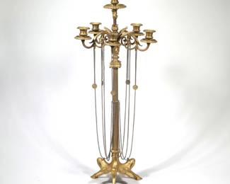 MONUMENTAL EMPIRE-STYLE GILT BRONZE CANDELABRUM | Gilt bronze floor candelabrum in an Empire or Neoclassical inspired form; vertical fluted standard rises from a tripartite base with carved foliate panels terminating in hoof feet, the upper section with scrolling foliate arms supporting a multi-light corona and a raised central finial; each candle socket sits above a beaded circular drip pan. Multiple fine metal chains suspend from the corona and shaft; chains are hung with round coin-form medallions pierced for attachment