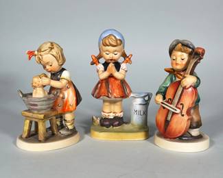(3pc) HUMMEL FIGURINES | Group of three hand-painted glazed porcelain figural children: a girl bathing a doll, a praying girl beside a milk can, and a boy playing a cello. Each is on its own oval or rectangular base; two bases show a blue printed mark and one base has a round black/gold sticker reading "Made in Japan." Colors are soft pastels and earth tones with handpainted detail and glossy glaze. Undersides show printed marks and a drilled hole in the center base.
