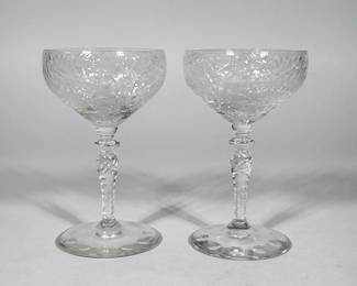 PAIR OF CUT GLASS CHAMPAGNE COUPES | Pair of clear cut-glass champagne coupes with shallow rounded bowls decorated with wheel-cut foliate and starburst motifs. Each glass is set on a knopped, spirally-cut stem with a small ring knop beneath the bowl and a circular foot with evenly spaced impressed discs. Clear, colorless glass with hand-cut style decoration.